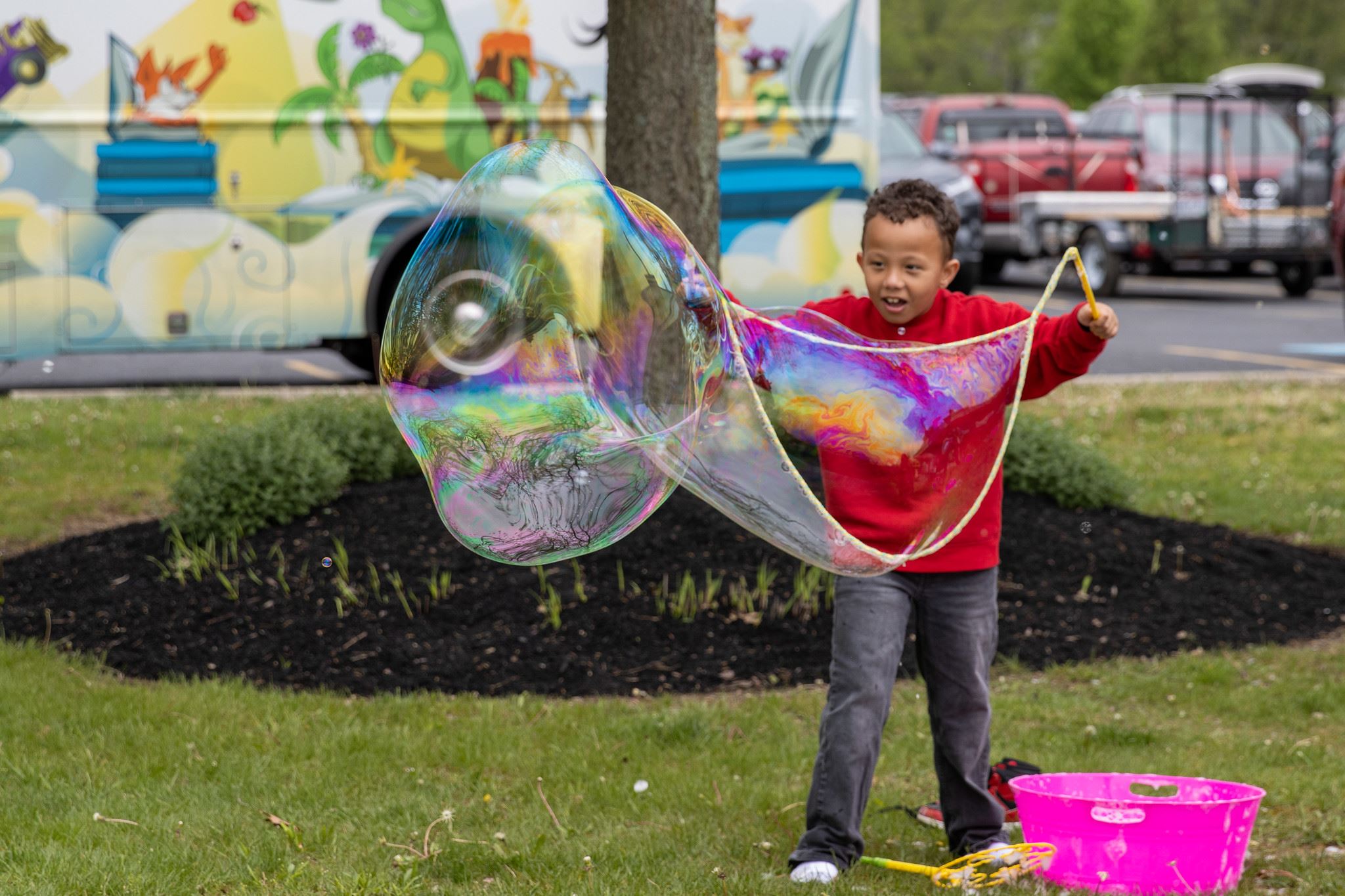 A young boy creates a giant rainbow-colored bubble using a wand at an outdoor event, smiling excited